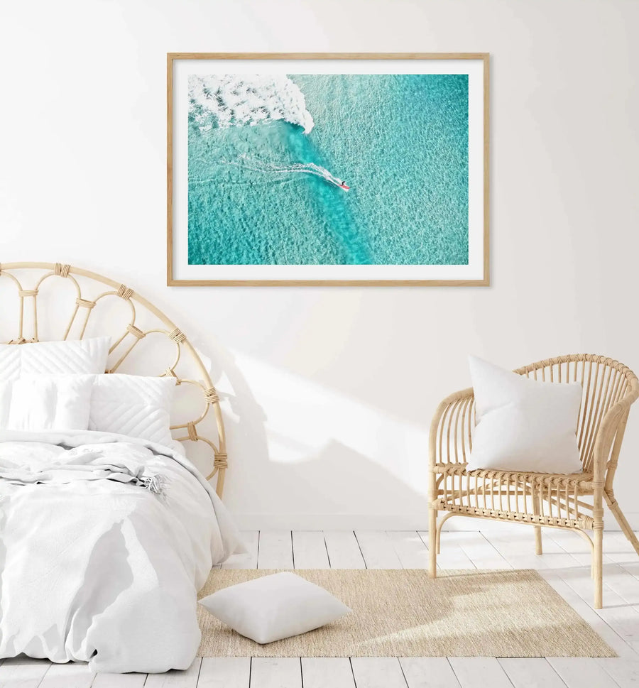 Aerial photograph of a surfer on a surfboard in turquoise ocean water near white breaking waves at Bondi Beach, displayed in a light wood frame with white border in a bright coastal bedroom with woven rattan bed and chair, white bedding, and pale timber flooring.