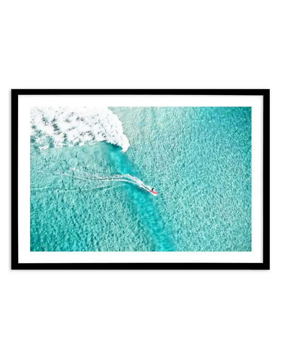 Aerial photograph framed in black showing a surfer in a red wetsuit paddling on a surfboard toward white-capped waves in turquoise ocean water at Bondi Beach, displayed as a wall art print with white border.