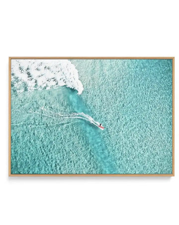 Framed aerial photograph of a surfer riding a wave in clear turquoise water, with a natural wood frame and white border. The composition captures the surfer on a red board, leaving a white foamy trail, with a large breaking wave to the left and clear, shallow water showing the sandy bottom.