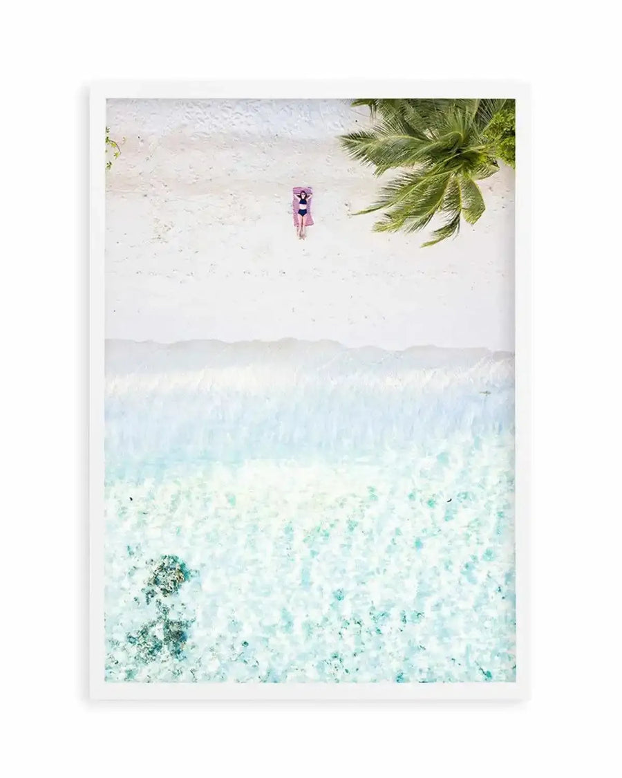Framed aerial photography art print of a tropical beach scene with a white frame and white border. The composition features a person in a dark swimsuit lying on a pink towel on white sand, with a palm tree in the upper right corner. Below the beach, gentle white waves meet clear turquoise water, revealing coral formations on the seabed.