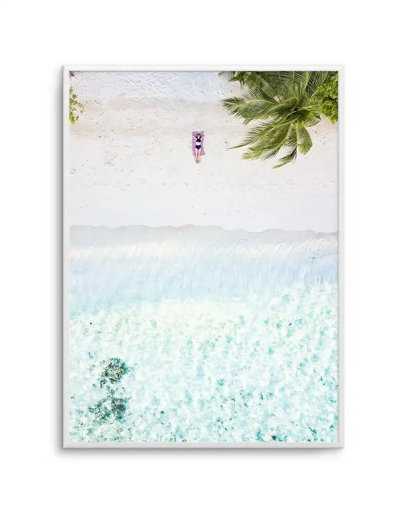 Framed aerial photograph of a tropical beach in the Maldives with a white frame and white border. The composition features a person lying on a pink towel on pristine white sand, with a palm tree casting a shadow on the right. Below the beach, gentle turquoise waves break on the shore, transitioning into clear, shallow ocean water with visible coral formations on the seabed.