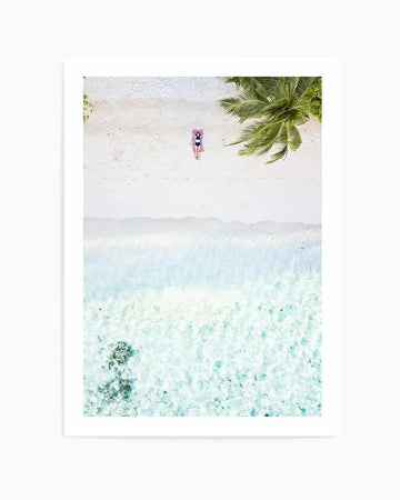 Aerial photography art print of a white sand beach and turquoise ocean. A person in a dark swimsuit lies on a pink towel on the sand, beneath the shade of a palm tree. The clear water reveals coral formations below the surface.