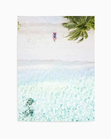 Aerial photography art print of a tropical beach scene with a person sunbathing on a pink and white striped towel on white sand, next to turquoise ocean water with gentle waves and a palm tree in the top right corner.