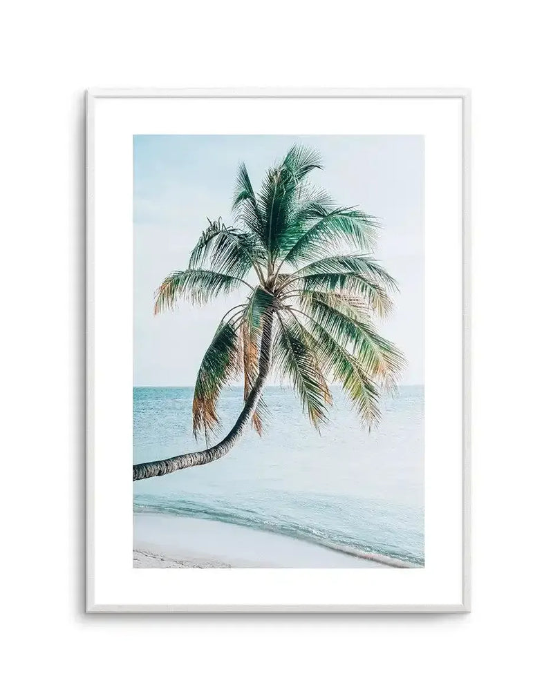 Framed art print featuring a photograph of a single palm tree with a curved trunk leaning toward the ocean. The tree has green fronds with some dried, tan-colored leaves at the base. Turquoise water and white sandy beach are visible in the background under a pale blue sky. The photograph is mounted in a white frame with a white border, creating a minimalist, coastal aesthetic.