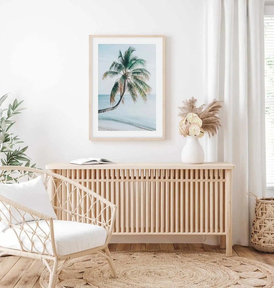 Framed art print featuring a tropical palm tree bending over calm turquoise ocean waters under a pale blue sky. The photograph-style image is mounted in a natural light wood frame with a white border. Displayed above a light wood slatted dresser in a bright, minimalist coastal-themed room with sheer white curtains, a woven rattan chair with white cushions, dried pampas grass in a white ceramic vase, potted green plant, and natural jute rug.