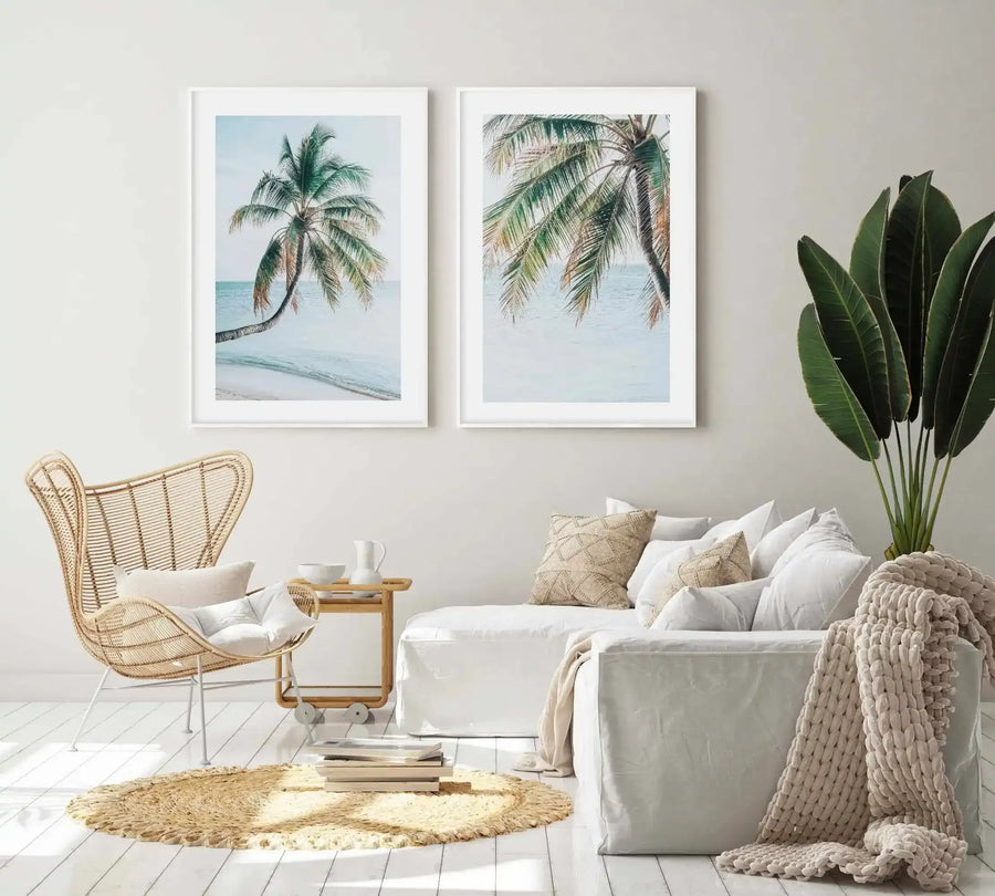 Framed art print featuring a watercolor illustration of a curved palm tree trunk extending into calm turquoise ocean waters under a pale blue sky. White wooden frame with white border. Displayed in a bright, minimalist bedroom setting alongside a woven rattan chair, neutral bedding, cream knit throw blanket, potted plant with long green leaves, and natural fiber round rug.