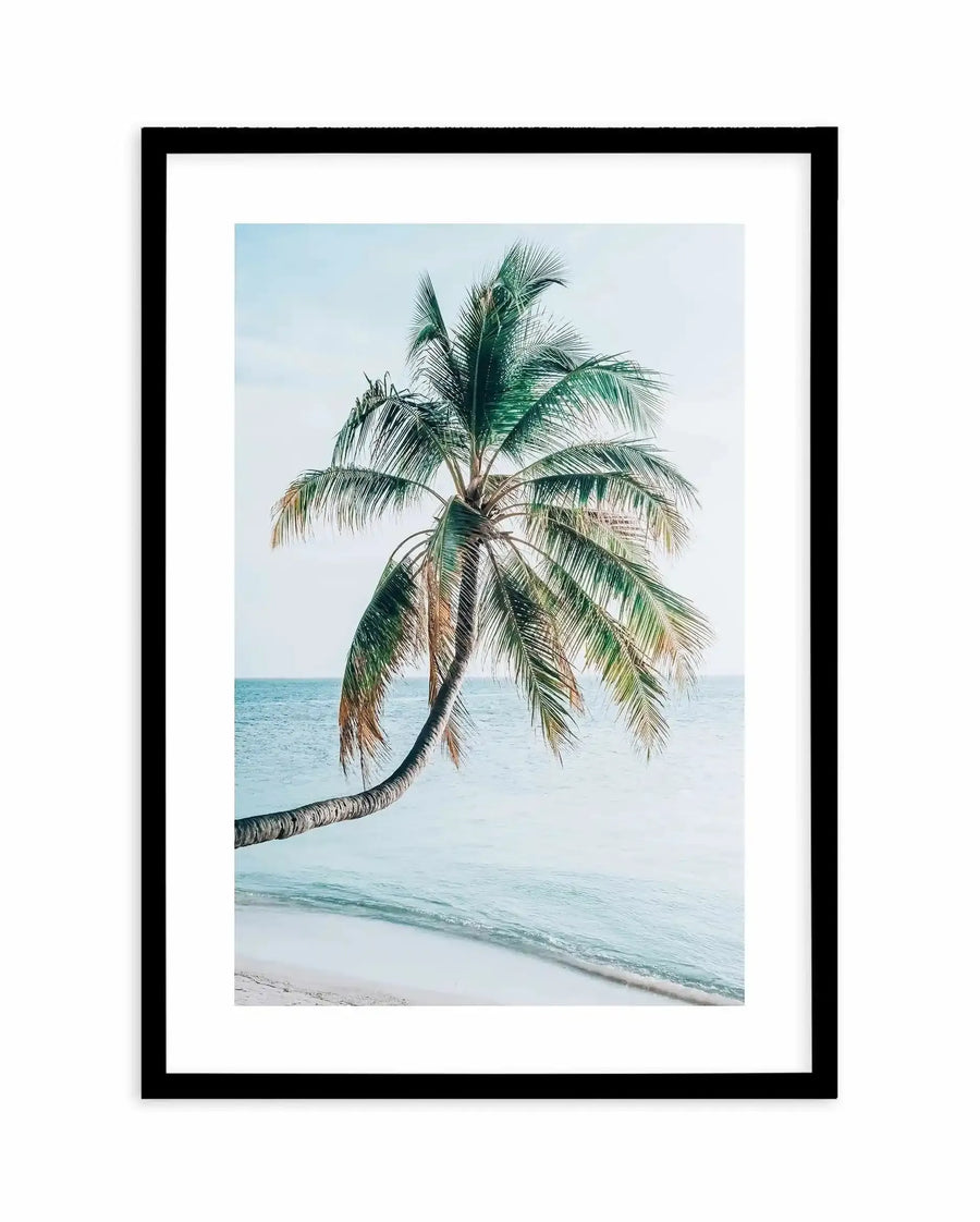 Framed art print featuring a photograph of a single palm tree with a curved trunk leaning over a sandy beach. The tree displays green and light tan fronds against a pale blue sky and calm turquoise ocean with gentle waves. The photograph has a soft, serene quality with muted colours. The print is matted with a white border and displayed in a black frame.