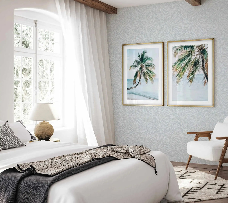 Maldivian Palm I Art Print displayed in a gold frame with white border on a light blue textured wall in a bright bedroom. The watercolor illustration depicts a palm tree with green fronds bending toward turquoise ocean water and sandy beach. Soft, muted colors include seafoam blue sky, cream sand, and natural green tones. The framed print hangs above a white bed with woven throw blankets in a serene coastal-inspired room with wooden ceiling beams and white curtains.