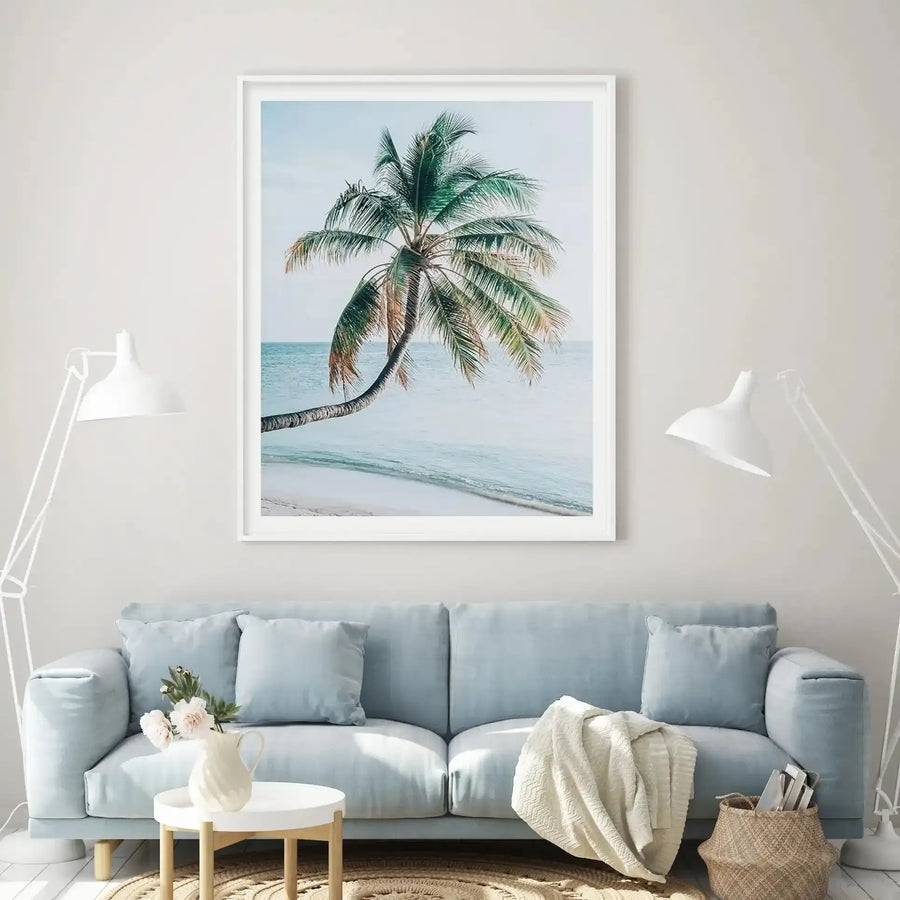 Framed art print featuring a photograph of a single palm tree with a curved trunk leaning toward turquoise ocean waters. The palm has green and golden-brown fronds against a pale blue sky and sandy beach. The image is displayed in a white frame with white border on a cream wall above a light blue-gray upholstered sofa styled with throw pillows, a cream blanket, white ceramic table lamp, and woven storage basket.