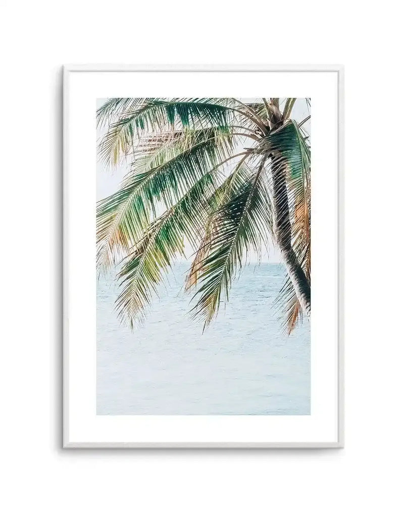 Framed art print featuring palm fronds hanging overhead against a soft turquoise ocean backdrop. The photograph captures delicate, feathery coconut palm leaves in shades of green, cream, and golden brown, rendered with fine detail and natural texture. The composition creates a serene tropical scene with the fronds positioned in the upper portion, allowing the calm, pale blue-green water to occupy the lower half. Presented in a light gray frame with white border.