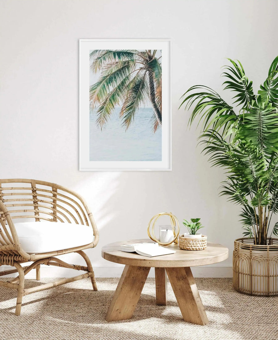 Framed art print titled Maldivian Palm II displayed on a white wall above a round wooden table. The print shows palm fronds with green and tan-colored leaves hanging downward against a pale blue sky and calm water backdrop, rendered in soft watercolor style. The artwork is presented in a white frame with white border. The room features natural wood furniture including a curved rattan chair with white cushion, woven plant baskets, and potted green plants, creating a tropical, coastal aesthetic.