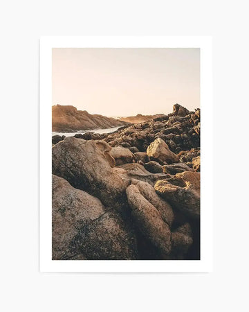 Framed photograph of a rocky coastal landscape at sunset with a natural wood frame and white border. The composition features a foreground of large, warm-toned boulders and rocks, with a body of water winding through a rocky inlet in the midground. The horizon shows a soft, hazy sky with the warm glow of the setting sun.