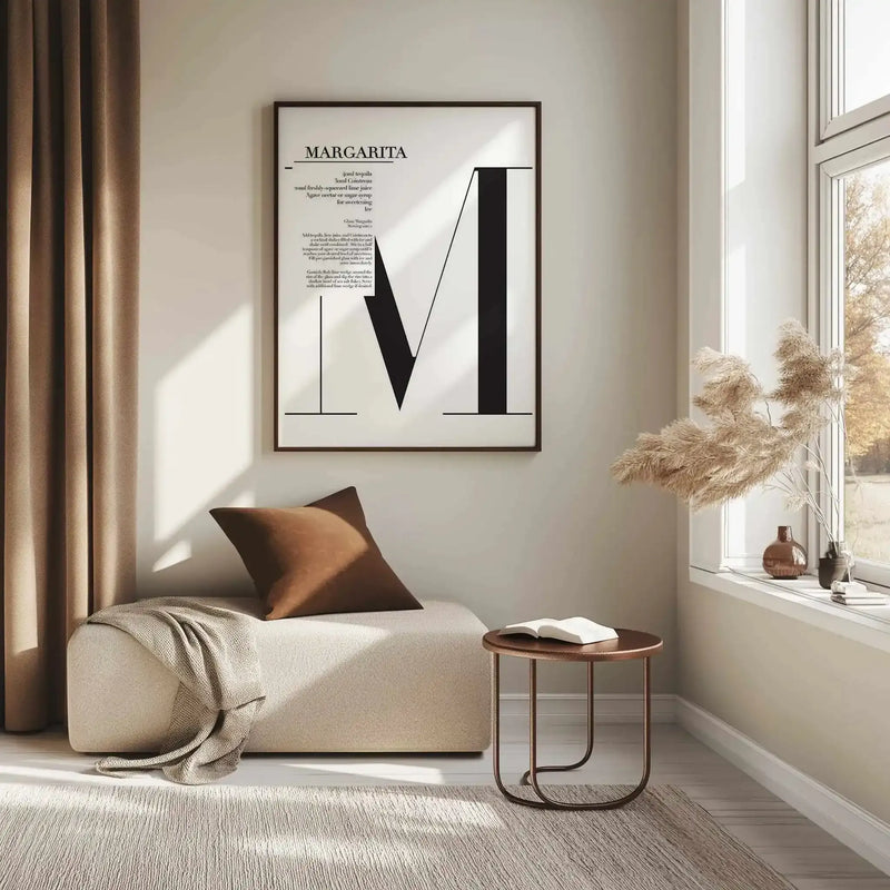 Margarita Art Print displayed in a black frame on a cream wall above a beige upholstered chaise lounge. The print features minimalist black geometric shapes forming an abstract letter M design on white paper, with recipe text at the top and additional descriptive text below. The framed artwork hangs in a bright, naturally-lit room with wooden side table, bronze cushion, cream throw blanket, and dried pampas grass in a vase on the windowsill.