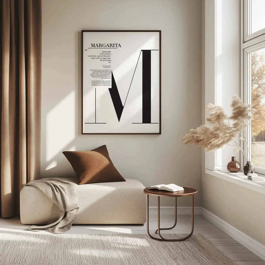 Margarita Art Print displayed in a black frame on a cream wall above a beige upholstered chaise lounge. The print features minimalist black geometric shapes forming an abstract letter M design on white paper, with recipe text at the top and additional descriptive text below. The framed artwork hangs in a bright, naturally-lit room with wooden side table, bronze cushion, cream throw blanket, and dried pampas grass in a vase on the windowsill.
