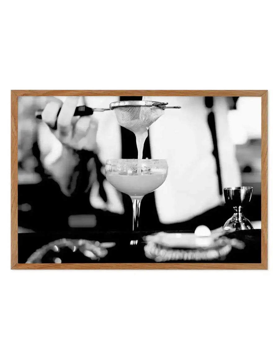 Black and white photograph of a bartender's hands straining a margarita cocktail through a fine mesh strainer into a coupe glass, with a white egg white foam top and ice visible. Wooden frame with white border. Vintage-style cocktail preparation scene with blurred background showing additional glassware and bar elements.