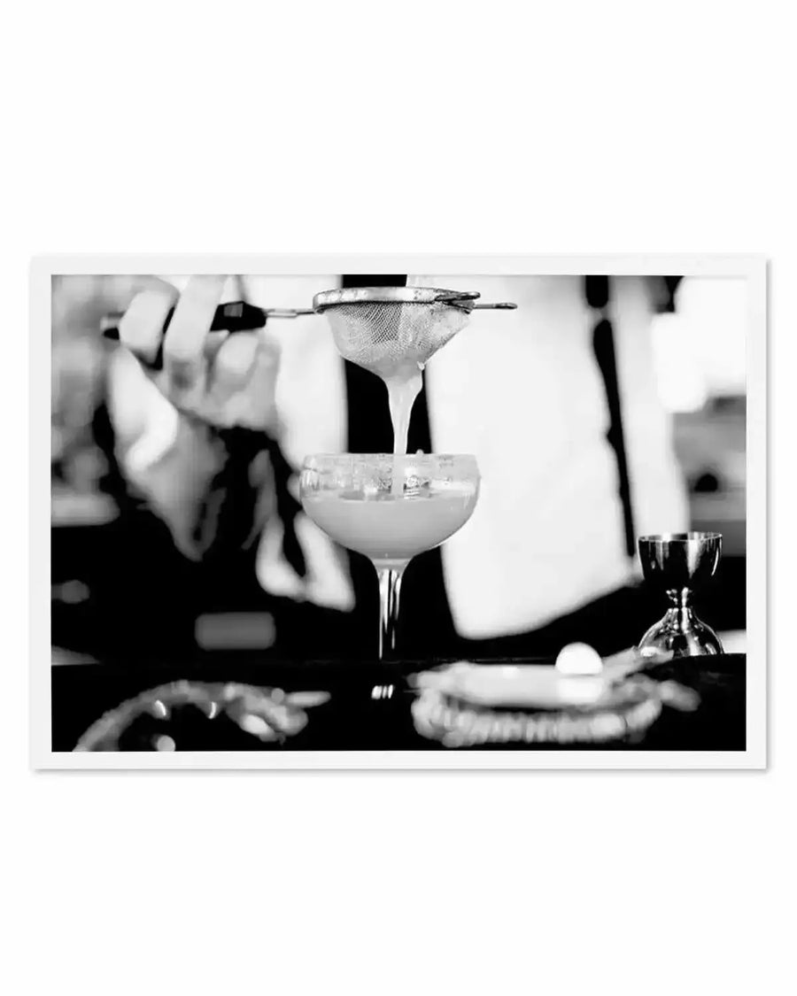 Black and white photograph showing a bartender's hands pouring a cocktail through a fine mesh strainer into a coupe glass filled with ice. The margarita drink is captured mid-pour with liquid flowing into the glass. A bar setup is visible with bottles and bar tools in soft focus in the background. The image has a vintage, classic aesthetic with high contrast and a white frame with white border.