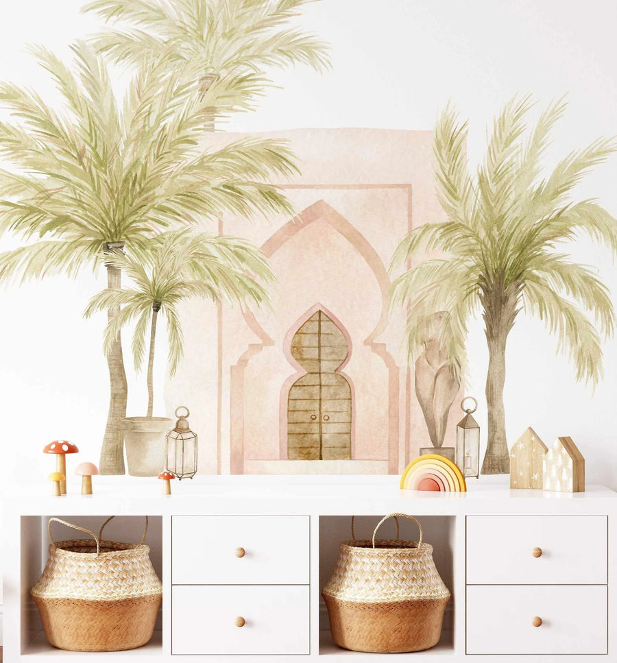 Lifestyle shot of a child's room featuring a Marrakech Palms Wallpaper Mural. The peel and stick wallpaper depicts a hand-painted scene of two large palm trees in muted sage green and olive tones flanking a pale pink Moroccan-style archway with a wooden door. The mural has a soft watercolour texture. Below the mural, a white storage unit holds two woven baskets and several decorative items, including wooden mushrooms, a rainbow toy, and small wooden houses.