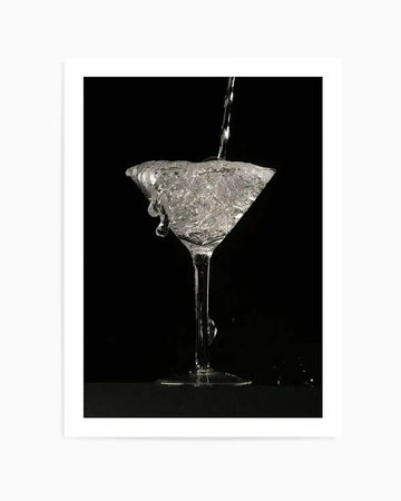 "Black and white photograph of a martini glass overflowing with clear liquid against a dark black background. A thin stream of liquid pours into the glass, causing it to spill over the rim, creating dynamic drips and splashes. The glass is illuminated, highlighting the texture and movement of the water."