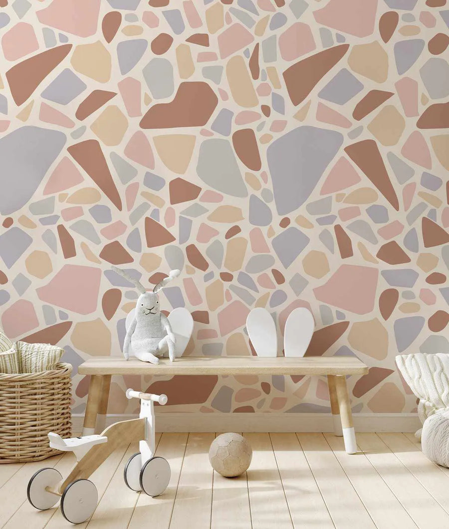 Nursery:
"Maya Terrazzo peel and stick wallpaper displayed in a child's room with a light wood bench, a woven basket, and toys. The wallpaper features a repeating pattern of irregular, organic shapes in various sizes and muted colours including dusty rose, terracotta, pale yellow, and light grey, set against a warm cream background. A white stuffed bunny with antlers sits on the bench next to two white bunny ear decorations. A small wooden balance bike and a textured ball are on the light wood floor."