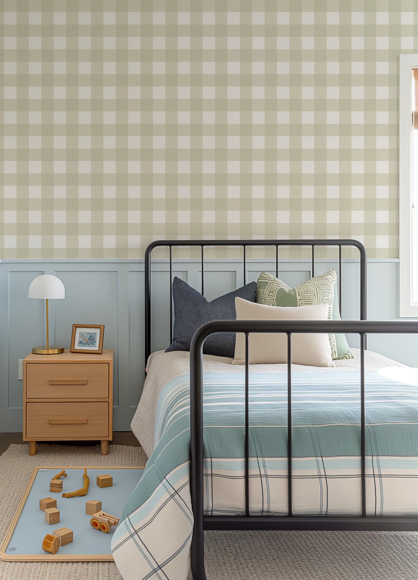 Lifestyle shot of a child's bedroom featuring a medium gingham check wallpaper in a new neutral green and soft white. The wallpaper covers the upper half of the wall, above light blue wainscoting. A black metal bed frame with a striped duvet in shades of blue, cream, and black is centered in the room. A light wood nightstand with a white and gold lamp and a framed picture sits to the left of the bed. Wooden toys are scattered on a light blue mat on the floor.