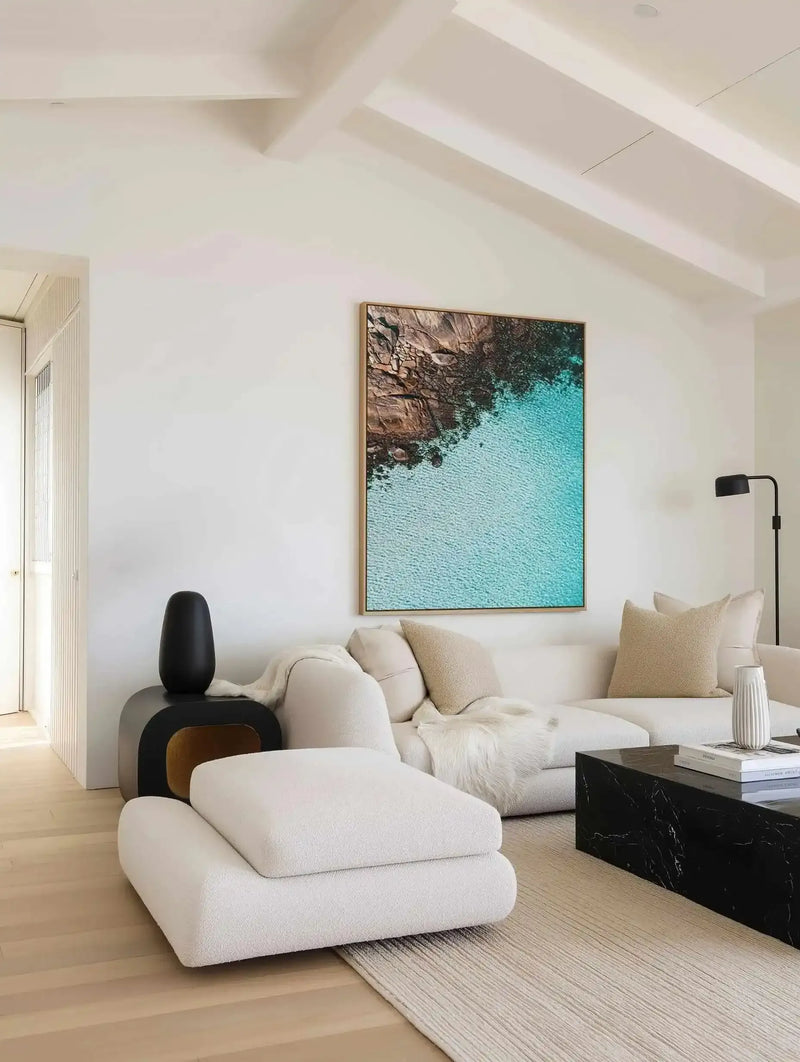 Aerial photograph framed in natural wood showing turquoise ocean water meeting rocky brown coastline, displayed above a cream upholstered sofa in a minimalist living room with light wood flooring, black accent furniture, and white vaulted ceiling.