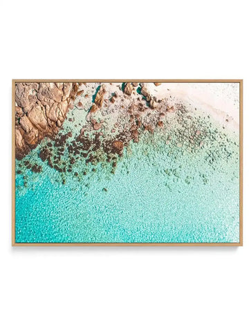 Aerial photograph framed in natural wood showing turquoise ocean water with rocky, tan and brown cliffs along the upper edge. The water transitions from bright cyan in the lower left to pale mint green near the sandy beach and rocks. Scattered dark boulders dot the shoreline. The natural wood frame has a warm honey tone finish with white border.