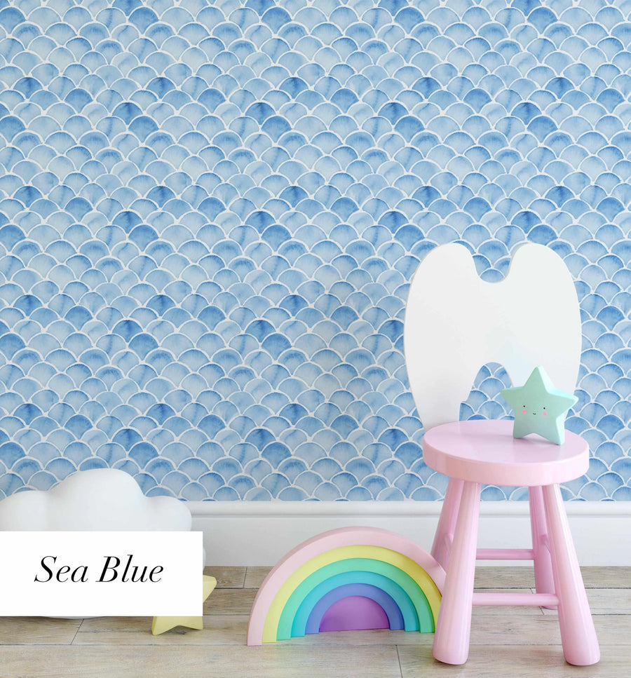 Lifestyle shot of a child's room featuring Sea Blue mermaid scales wallpaper. The wallpaper has a repeating pattern of hand-painted scallop shapes in varying shades of light to medium blue with visible watercolor brushstrokes and white outlines, creating a soft, ocean-inspired texture. The room includes a light pink stool, a white cloud-shaped object, a pastel rainbow toy, and a mint green star-shaped light with a smiling face.