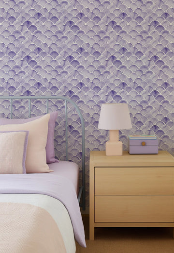 "Peel and stick wallpaper featuring a repeating pattern of hand-painted mermaid scales in varying shades of lavender and deep purple on a soft white background. The scalloped design has a loose, organic watercolour quality with visible brushstroke texture."