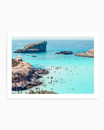Framed photograph of a vibrant, crowded beach scene in Malta with a white frame and border. The composition features turquoise blue water filled with swimmers, surrounded by rocky cliffs where people are sunbathing and relaxing under blue umbrellas. Several boats are visible in the distance, and a large rock formation rises from the sea under a clear blue sky.