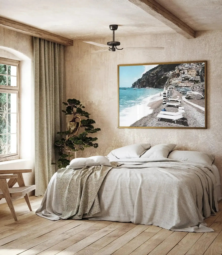Framed art print titled "Midday in Positano" displayed above a linen bed in a minimalist bedroom. The photograph shows a Mediterranean beach scene with turquoise waters, white sandy shore, striped beach loungers, and white-washed buildings cascading up a clifside. The print has a natural wood frame and is mounted on a textured beige wall. The bedroom features cream-colored bedding, a woven throw blanket, wooden flooring, a bonsai tree in a pot, flowing cream curtains, and a black ceiling fan.