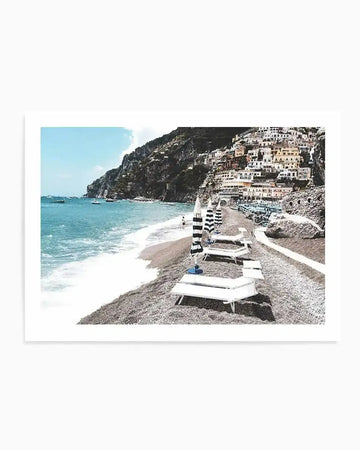 "Photography art print of a vibrant coastal town nestled into a hillside overlooking a beach, printed on fine art paper. The composition features a pebbled beach with white sun loungers and striped umbrellas in the foreground, leading to turquoise ocean waves and boats, with a town of multi-coloured buildings climbing the steep green hillside under a bright blue sky."