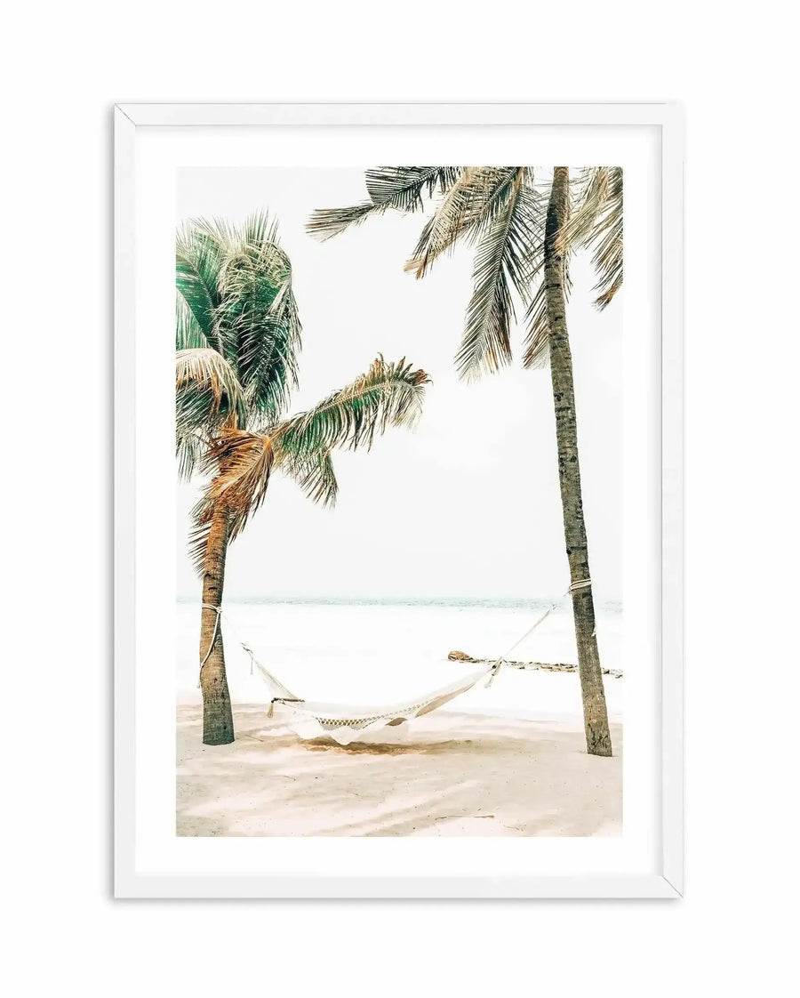 Framed art print featuring a tropical beach scene with two palm tree trunks in the foreground flanking a white hammock on sandy beach, calm turquoise ocean visible in the background under a pale sky. Palm fronds with green and golden-brown tones hang from above. The photograph has a soft, muted color palette with warm sand tones and natural lighting. White wooden frame with white border surrounding the artwork.