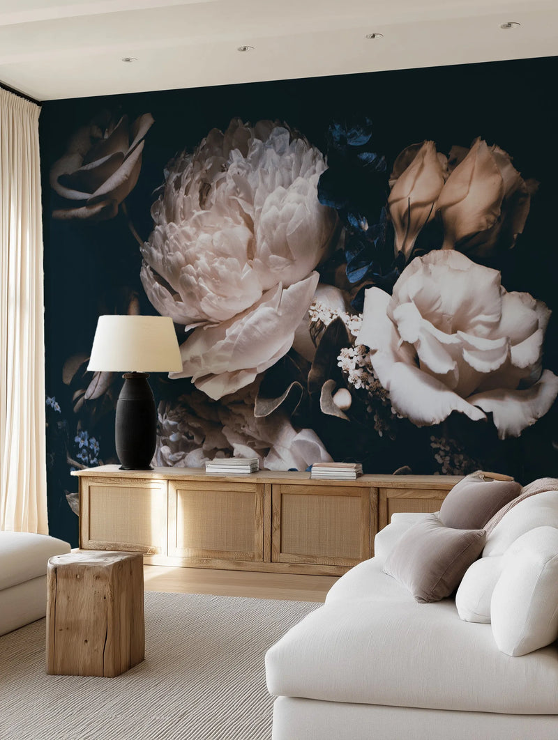 Peel and stick wallpaper mural featuring a large-scale botanical design of oversized, dark and moody flowers in shades of cream, blush, and deep blue on a dark navy background, displayed on a living room wall. The wallpaper is behind a light wood console table with woven cane doors, topped with a black lamp and stacks of books. A white sofa with taupe and white pillows, and a light wood side table sit on a textured cream rug in the foreground.