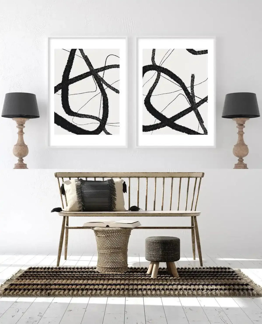 Two framed abstract art prints displayed side-by-side on a white wall above a light wood bench in a minimalist living room. Each print features thick, bold, and thin, delicate black lines forming an abstract, intertwined pattern on a white background, with white frames and white borders. The room includes two black-shaded table lamps with carved wood bases, a striped brown and black rug, a woven stool, and a small dark grey upholstered stool.