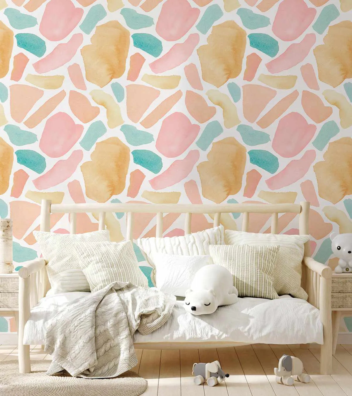 Lifestyle shot of a child's bedroom featuring a daybed with white bedding and pillows against a wallpapered wall. The wallpaper features a repeating pattern of abstract, organic shapes in soft pastel shades of blush pink, peach, light yellow, and teal, with a watercolor texture on a white background. A white polar bear plush toy rests on the daybed, and two small elephant toys are on the light wood floor.