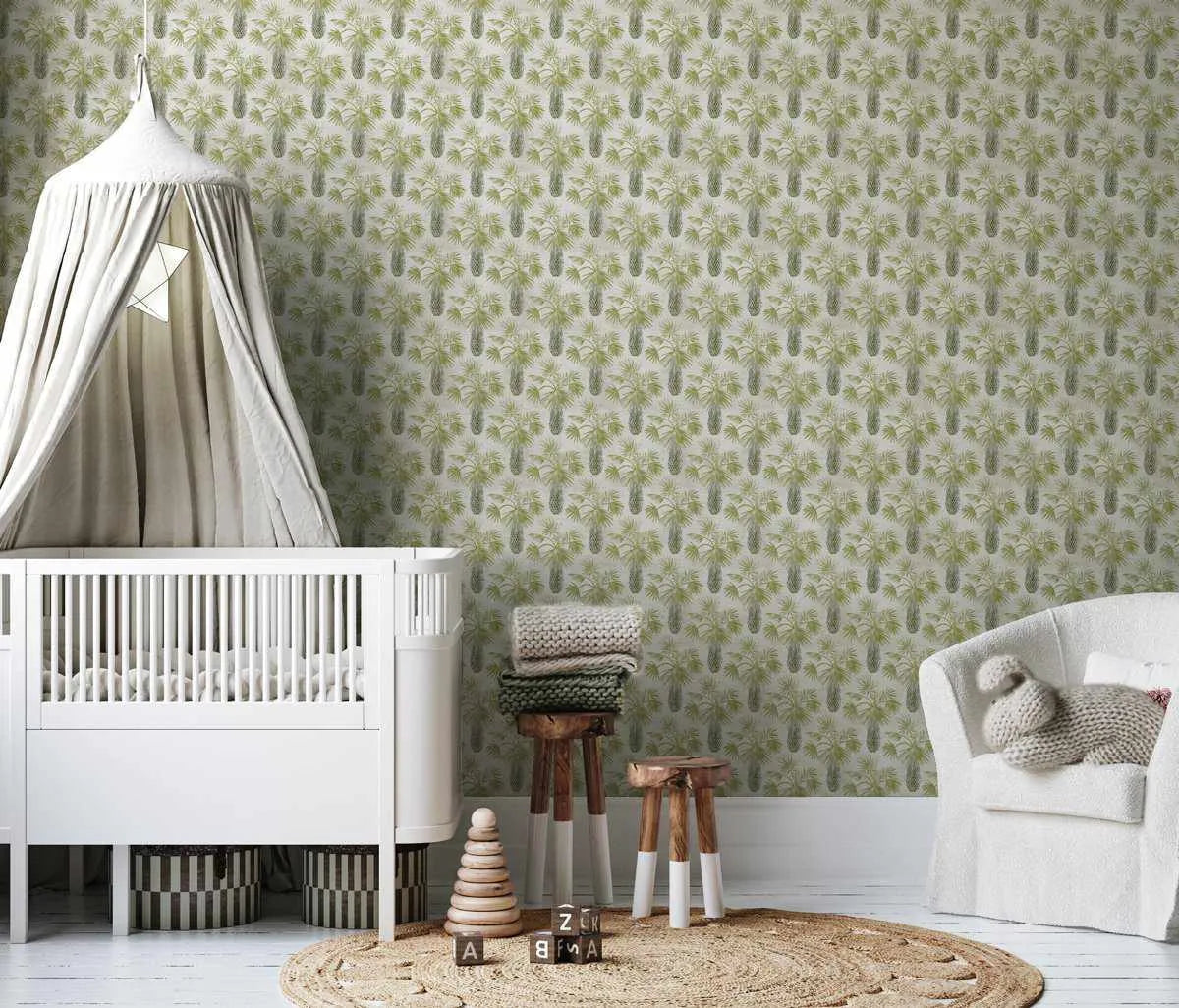 Mimosa Wallpaper displayed on a nursery room wall above white wainscoting. The wallpaper features a repeating pattern of soft sage green mimosa flower clusters and elongated leaf shapes on a cream background. The room includes a white crib with canopy, white armchair, wooden stools with knitted blankets, a round woven rug, and wooden toys, creating a calm, nature-inspired aesthetic. The wallpaper has a subtle, botanical illustration style with muted earth tones.