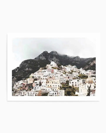 "Photography art print of Positano, Italy, featuring a hillside town with numerous white and pastel-coloured buildings cascading down a steep mountain, partially obscured by a soft mist or low clouds. The composition captures the dense cluster of houses with terracotta roofs and a prominent dome-shaped church roof in the lower right, set against a dramatic, dark mountain range under an overcast sky."