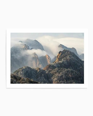 "Framed photograph of the misty Huangshan Mountain in China, with a white border. The composition features multiple mountain peaks shrouded in soft, ethereal clouds and mist, with the lower slopes covered in snow-dusted trees and foliage. The warm light of the sun illuminates some of the mountain faces, creating a serene and majestic atmosphere."