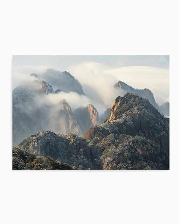 Photography art print featuring a majestic mountain range shrouded in mist and clouds, with peaks emerging into sunlight. The mountains are covered in snow-dusted trees and foliage, with warm golden light illuminating some of the rocky faces. The composition captures a serene and ethereal mood with soft light and atmospheric perspective.