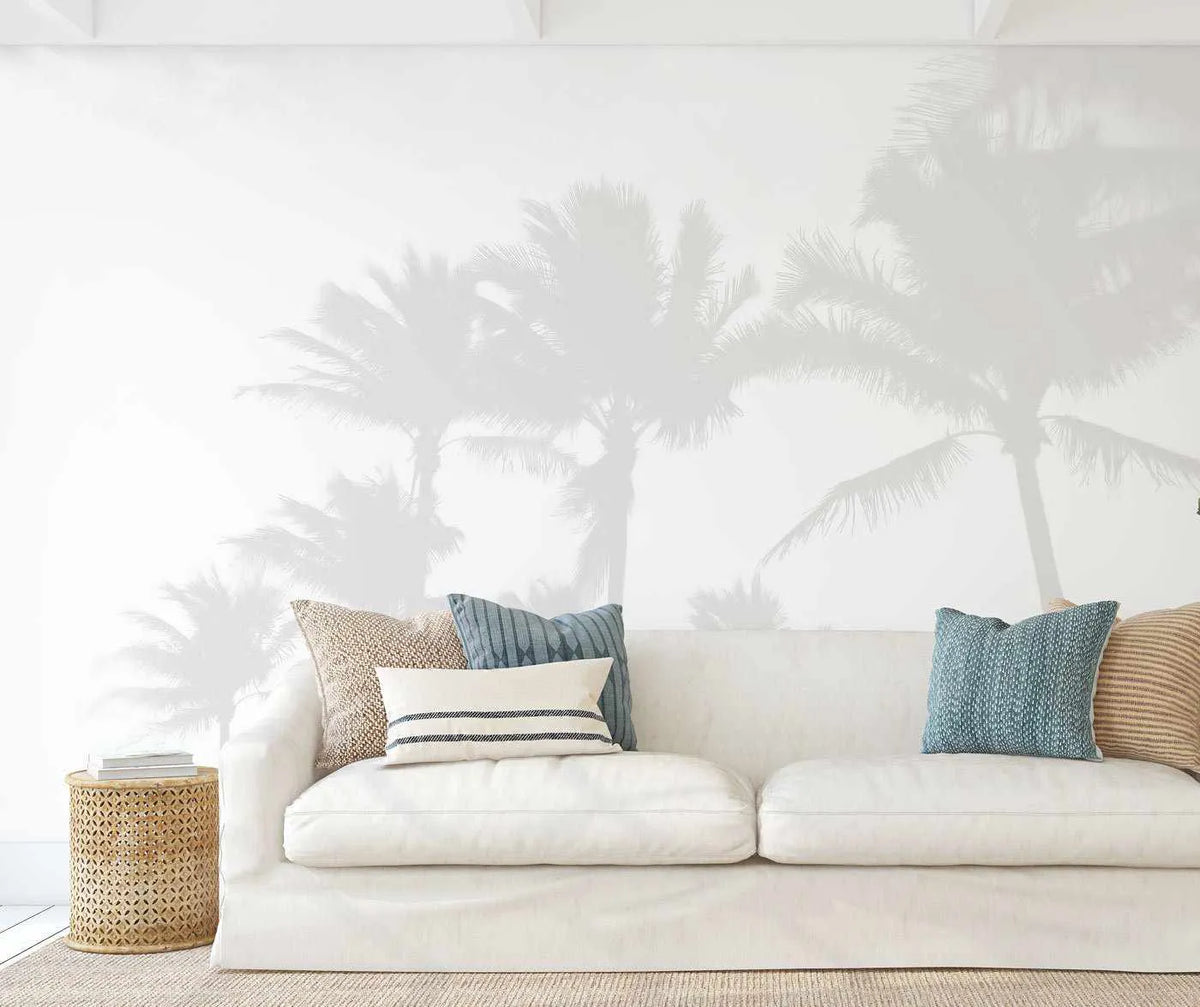 "Misty Palms peel and stick wallpaper mural displayed on a white wall above a white slipcover sofa in a bright living room. The wallpaper features a soft, ethereal design of palm tree silhouettes in pale grey against a white background, creating a serene, tropical ambiance. The sofa is adorned with textured cushions in natural beige, striped navy and white, and solid teal blue. A woven rattan side table with books sits to the left of the sofa, and a light-coloured rug covers the floor."