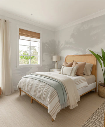 "Bedroom with a bed featuring a rattan headboard, white and beige striped duvet, and a light blue throw blanket. The walls are covered in a peel and stick wallpaper featuring a repeating pattern of palm tree shadows in a soft grey on a light cream background, creating a serene, tropical ambiance. A window with a bamboo blind and white curtains looks out onto lush greenery. A white nightstand with a ceramic lamp sits beside the bed, and a large green plant in a woven basket is on the floor."