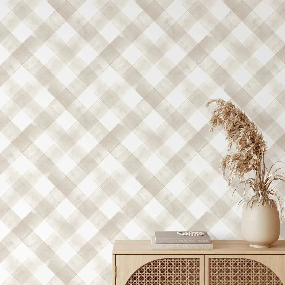 "Modern Check in Caramel Wallpaper applied to a wall above a light wood cabinet with woven cane doors. The wallpaper features a repeating pattern of hand-painted diagonal checks in warm caramel and soft white, with visible brushstroke texture. A vase of dried pampas grass and a stack of books sit on the cabinet."