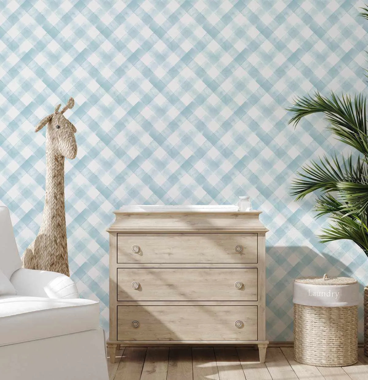 Lifestyle shot of a nursery featuring Modern Check in Blue Salt Lake Wallpaper. The wallpaper features a repeating pattern of hand-painted diagonal checks in a soft, muted blue and white, creating a watercolor-like texture. The room includes a light wood changing table with three drawers, a white armchair, a woven giraffe toy, a green leafy plant, and a woven laundry basket with a white fabric liner.