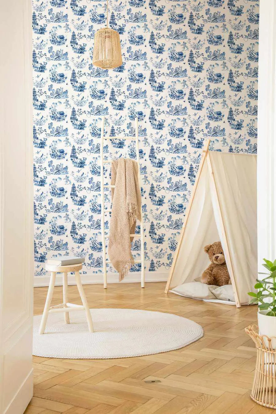 Blue and white toile wallpaper covering an entire bedroom wall, featuring an intricate repeating pattern of pastoral landscapes with trees, buildings, figures, and decorative flourishes in cobalt blue on a cream background. The wallpaper is displayed in a styled children's room with natural wood flooring, a white wooden tent structure containing a brown teddy bear, a woven pendant lamp, a white stool with a beige throw blanket, and a potted green plant, creating a serene, classic aesthetic.