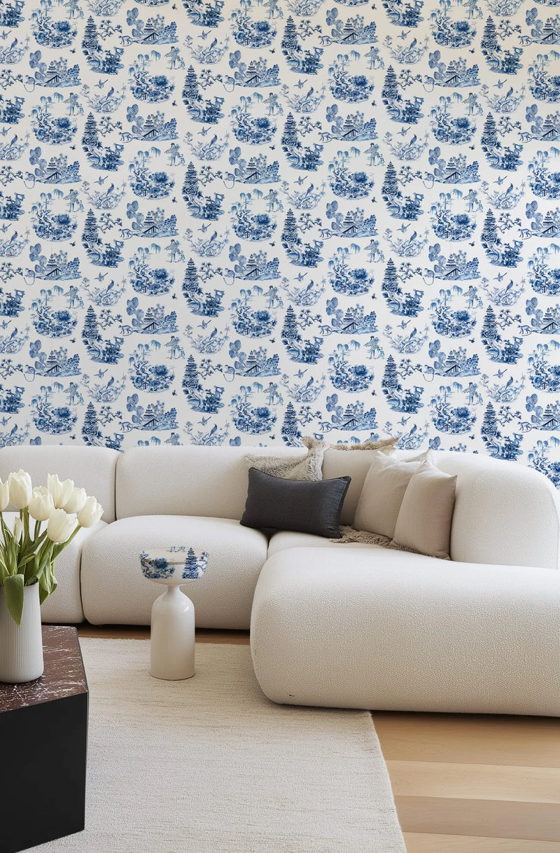 Blue and white toile wallpaper with repeating pastoral scenes featuring pagodas, weeping willow trees, bridges, and figures in an Asian-inspired landscape design displayed on a living room wall behind a curved cream-colored sectional sofa with decorative pillows and a white ceramic vase with white tulips on a light wooden floor.