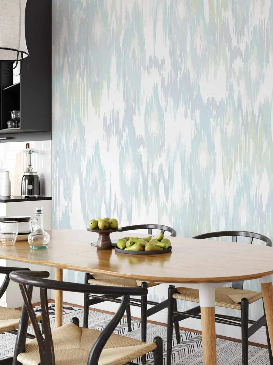 Lifestyle shot of a dining room featuring a wall covered in Molten Wave wallpaper. The wallpaper has a repeating abstract pattern of vertical, jagged brushstrokes in shades of pale blue, light grey, and soft sage green on a white background, creating a fluid, ikat-inspired design. A light wood oval dining table with black chairs and woven seats is in the foreground, adorned with two bowls of green pears. A black kitchen cabinet and white tiled backsplash are visible on the left.