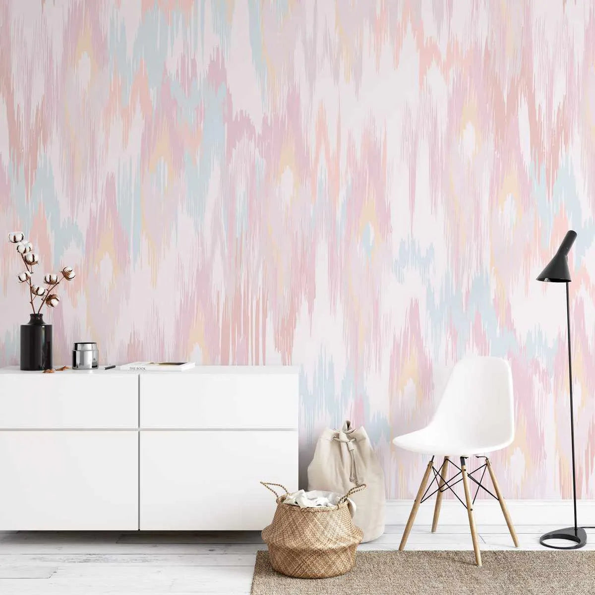 Molten Wave wallpaper featuring a repeating pattern of vertical, irregular brushstrokes in soft pastel shades of blush pink, pale blue, light yellow, and cream, creating a textured, abstract design. The wallpaper is displayed on a wall in a minimalist living room with a white low-profile cabinet, a black floor lamp, a white modern chair with wooden legs, and a woven basket on a jute rug.