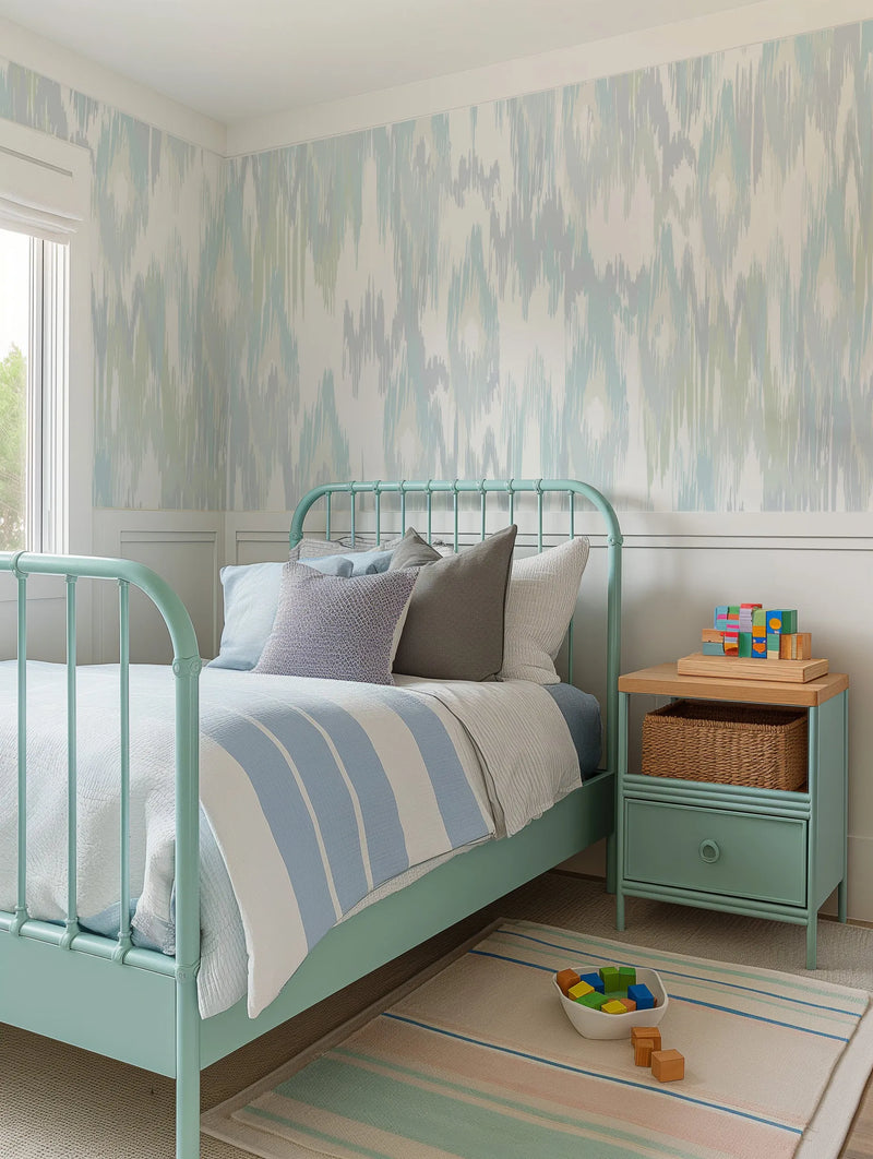 Lifestyle shot of a child's bedroom featuring Molten Wave wallpaper in a light blue and sage green abstract pattern. The wallpaper covers the upper half of the walls, above white wainscoting. A mint green metal bed with a white and blue striped duvet and various pillows is positioned against the wall. A matching mint green nightstand with a wooden top, a woven basket, and building blocks sits beside the bed. A striped rug in muted pink, green, and blue lies on the floor.