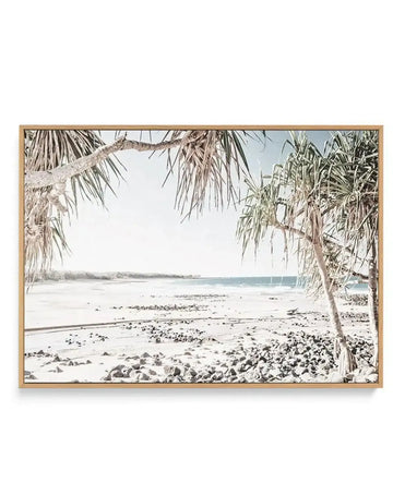 Framed canvas art print featuring a serene coastal beach scene with a soft, muted colour palette, displayed in a natural oak frame. The composition captures a wide expanse of light-coloured sand and small grey rocks leading to gentle blue ocean waves under a bright, slightly overcast sky. Two pandanus trees with long, spiky leaves frame the scene from the left and right foreground, their branches extending into the upper portion of the image.