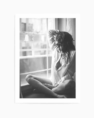 Framed black and white photograph of a woman sitting on a window sill, looking out of the window. She is wearing an unbuttoned white shirt and has long, dark, wavy hair. Her legs are crossed, and one arm is raised, resting on her head. The window frames a blurred view of buildings outside.